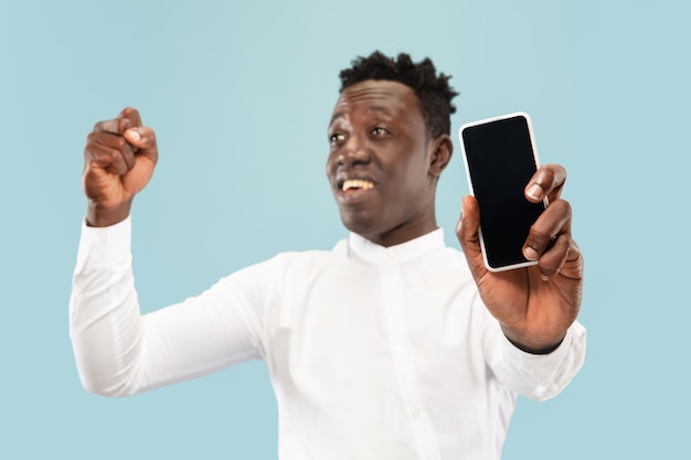 Молодой афро-американский мужчина позирует со смартфоном, изолированным на синем фоне студии, выражение лица. Красивый мужской портрет половинной длины. Понятие о человеческих эмоциях, выражении лица.