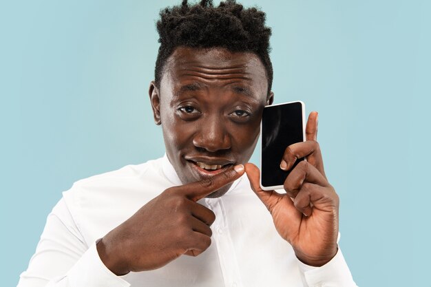 Young african-american man posing with smartphone isolated on blue studio