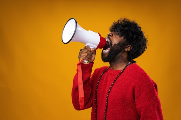 Young african american man in red hoodie screaming in megaphone against yellow background