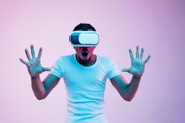 Young african-american man's playing in VR-glasses in neon light on gradient background. Male portrait