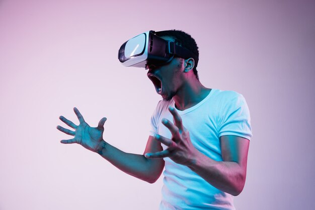 Молодой афро-американский мужчина играет в VR-очках в неоновом свете на градиентном фоне. Мужской портрет