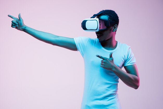 Young african-american man's pointing and using VR-glasses in neon light on gradient