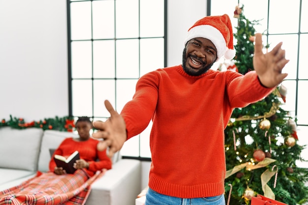 Молодой афроамериканец, стоящий у рождественской елки, смотрит в камеру, улыбаясь с распростертыми объятиями для объятий, веселое выражение лица, обнимающее счастье
