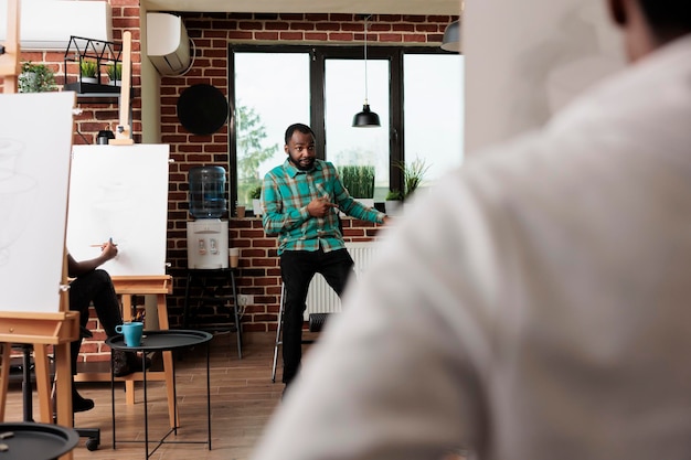 Free photo young african american man teaching art to people in creative studio, male instructor demonstrating sketching techniques to students sitting at easels during group drawing workshop