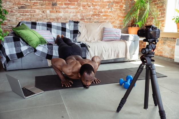 Free photo young african-american man teaching at home online courses of fitness, aerobic, sporty lifestyle during quarantine, reording on camera, streaming