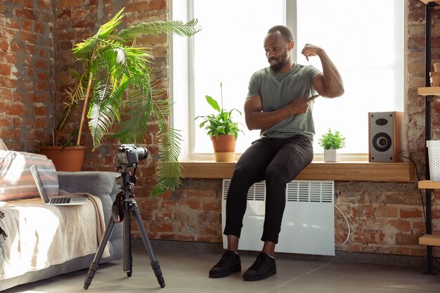 Young african-american man teaching at home online courses of fitness, aerobic, sporty lifestyle during quarantine, reording on camera, streaming