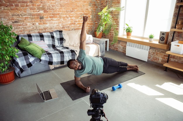 Young african-american man teaching at home online courses of fitness, aerobic, sporty lifestyle during quarantine, reording on camera, streaming