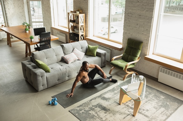 Young african-american man training at home , doing exercises of fitness, aerobic. 