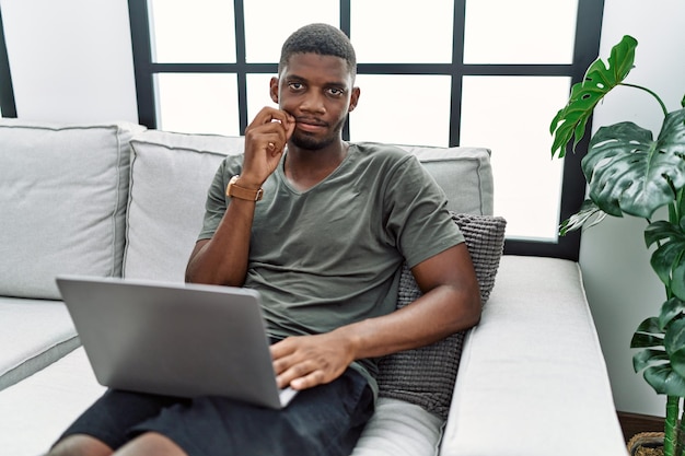 無料写真 自宅でラップトップを使用している若いアフリカ系アメリカ人男性がソファに座り、口と唇を指でジッパーのように閉じます。秘密で沈黙、タブーな話