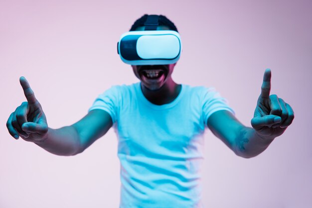 Young african-american man in VR-glasses in neon light on gradient background. Male portrait