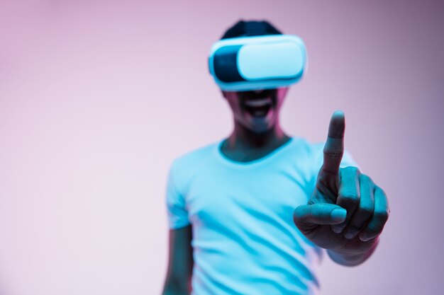 Young african-american man in VR-glasses in neon light on gradient background. Male portrait