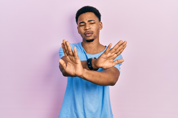 Молодой афроамериканец в повседневной синей футболке с выражением отказа скрещивает руки и ладони, делая отрицательный знак злое лицо