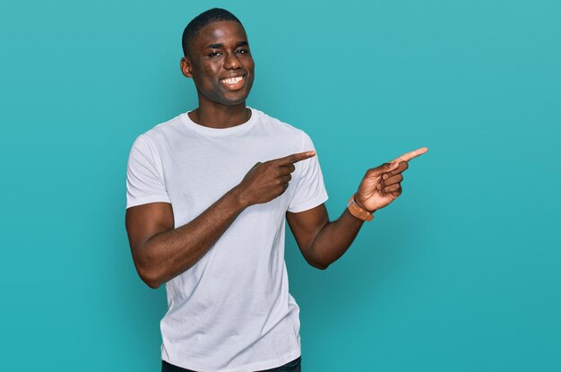 Молодой афроамериканец в повседневной белой футболке улыбается и смотрит в камеру, указывая двумя руками и пальцами в сторону