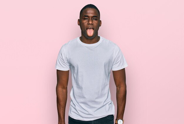 Молодой афроамериканец в повседневной белой футболке с высунутым языком, довольный забавным выражением лица. концепция эмоций.