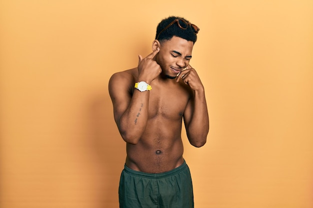 Free photo young african american man wearing swimwear and swimmer glasses covering ears with fingers with annoyed expression for the noise of loud music deaf concept