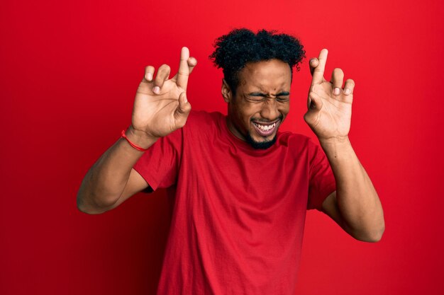 Молодой афроамериканец с бородой в повседневной красной футболке жестикулирует скрещенными пальцами, улыбаясь с надеждой и закрытыми глазами удачи и суеверной концепции