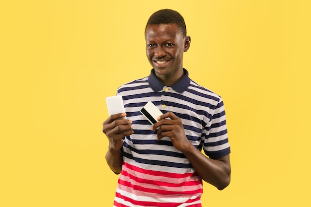 Молодой афро-американский человек со смартфоном и дебетовой картой, изолированные на желтом фоне студии