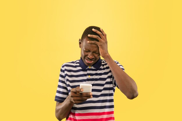 黄色のスタジオの背景、顔の表情に分離されたスマートフォンを持つ若いアフリカ系アメリカ人の男。美しい男性の半分の長さの肖像画。人間の感情、顔の表情の概念。