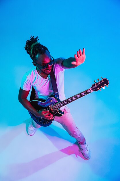 Young african-american musician playing the guitar like a rockstar on blue  background in neon light.