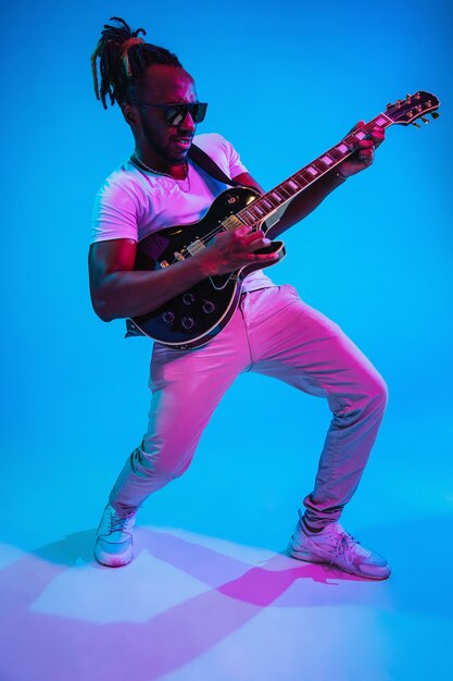 Young african-american musician playing the guitar like a rockstar on blue wall in neon light. 