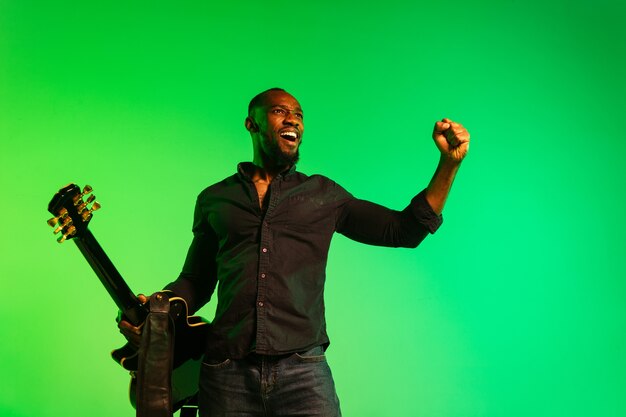 Young african-american musician playing the guitar like a rockstar on gradient green-yellow background. Concept of music, hobby, festival, open-air. Joyful  guy improvising, singing song.