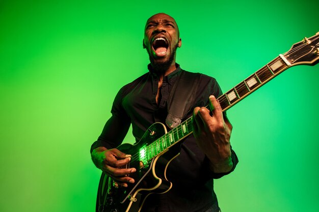 緑黄色のグラデーションの背景でロックスターのようにギターを弾く若いアフリカ系アメリカ人のミュージシャン。
