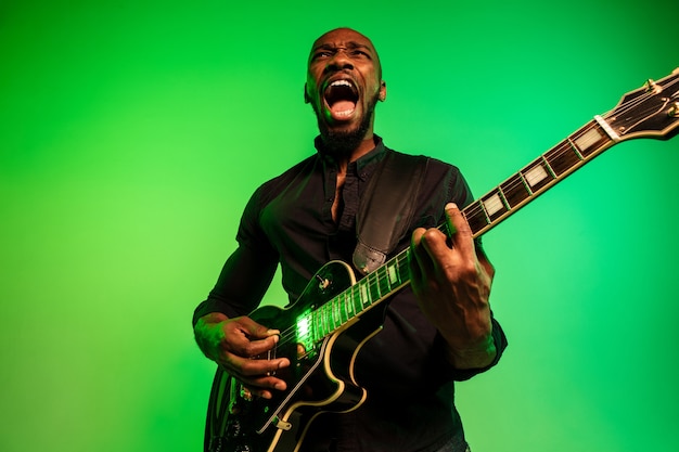 그라데이션 녹색 노란색 배경에 록 스타처럼 기타를 연주하는 젊은 아프리카 계 미국인 음악가.