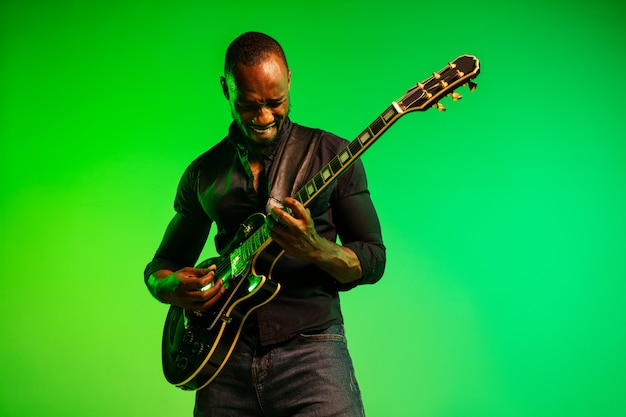 Young african-american musician playing the guitar like a rockstar on gradient green-yellow background.