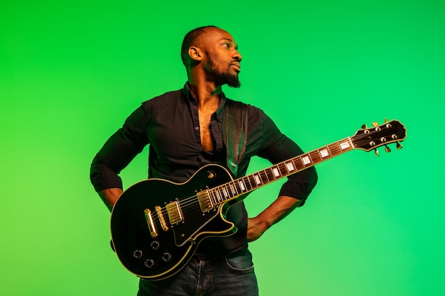 Young african-american musician playing the guitar like a rockstar on gradient green-yellow