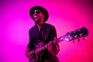 Young african-american musician playing the guitar like a rockstar on gradient purple-pink background in neon light. concept of music, hobby. joyful  guy improvising and singing a song.