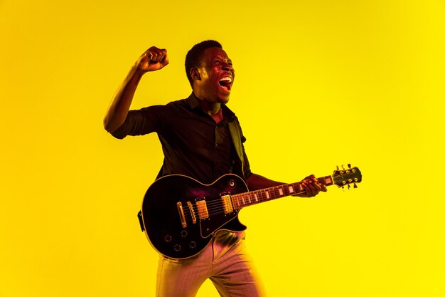 Young african-american musician playing the guitar like a rockstar on yellow background in neon light.