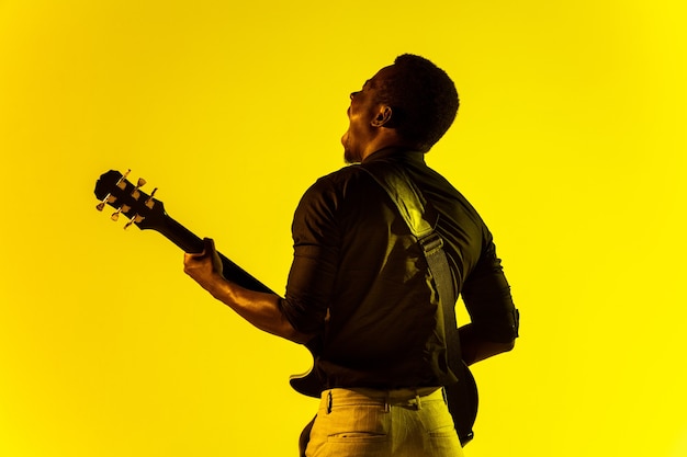 ネオンの光の中で黄色の背景にロックスターのようにギターを弾く若いアフリカ系アメリカ人のミュージシャン。