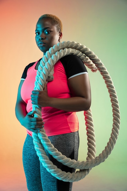 Young african-american plus size female model's training on gradient wall in neon light.