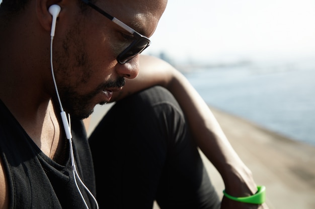 Free photo young african american rapper in black top listening to new tracks outside under blue sky. handsome and serious man sitting alone on the roadside and chatting with his friends on his digital device.