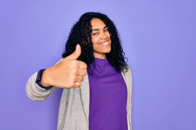 Giovane donna sportiva afroamericana che indossa una felpa casual su sfondo viola facendo il pollice felice in alto gesto con la mano approvando l'espressione guardando la telecamera mostrando successo