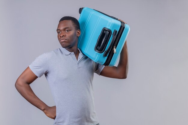 Young african american traveler man in grey polo shirt holding blue suitcase looking aside displeased and frowning