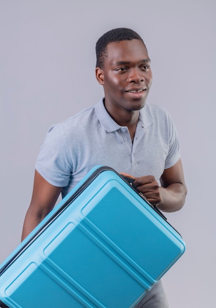 Young african american traveler man in grey polo shirt holding blue suitcase looking aside smiling