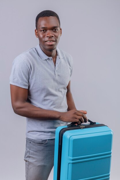 青いスーツケースを前向きで幸せな笑顔を保持している灰色のポロシャツの若いアフリカ系アメリカ人旅行者の男