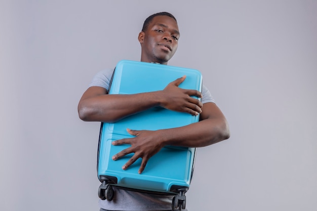 Young african american traveler man in grey polo shirt hugging his blue suitcase with confident smile on face