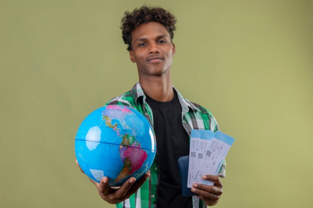 Young african american traveler man holding air tickets and globe stretching it out to camera looking confident smiling standing over green background