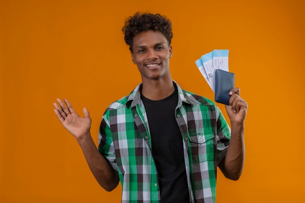Молодой афро-американский путешественник мужчина держит авиабилеты, весело улыбаясь и счастливо глядя в камеру, стоящую на оранжевом фоне