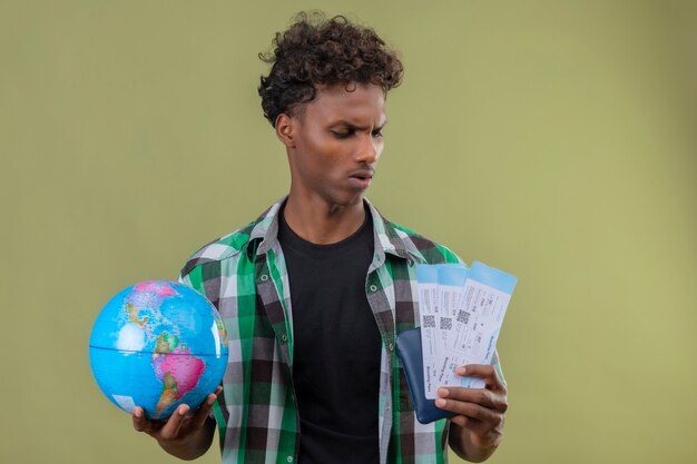 Young african american traveler man holding globe and air tickets looking at them with serious expression on face frowning standing over green background