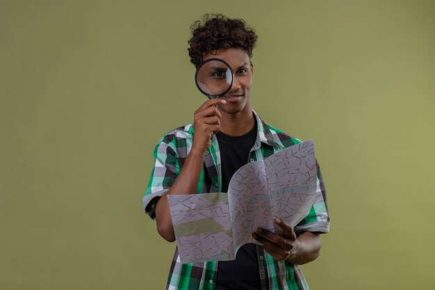 Young african american traveler man holding map looking at camera through magnifying glass smiling standing over green background