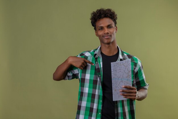 緑の背景の上に立って自信を持って笑顔でカメラを見て自分を指しているマップを保持している若いアフリカ系アメリカ人旅行者男