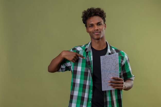 Young african american traveler man holding map pointing to himself looking at camera with confident smile standing over green background