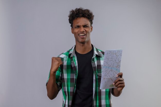 Young african american traveler man holding map raising fist exited and happy raising fist rejoicing his success standing over white background