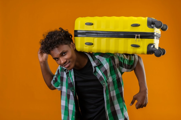 Young african american traveler man holding suitcase on his back looking at camera positive smiling standing over orange background