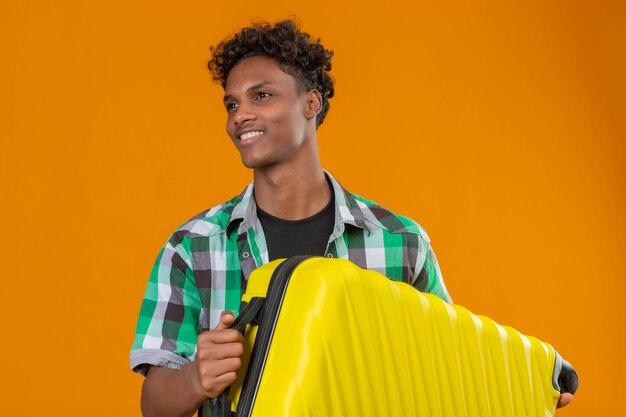 Young african american traveler man holding suitcase looking aside smiling happy and positive standing over orange background