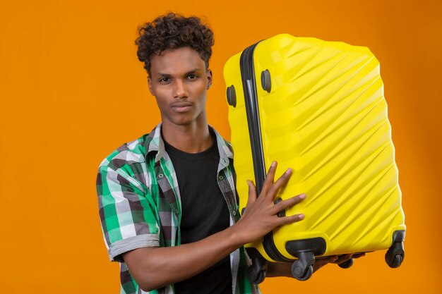 Young african american traveler man holding suitcase looking at camera with confident serious expression on face standing over orange background