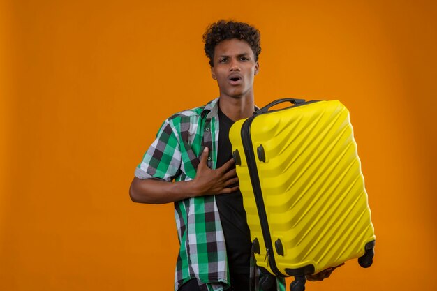 オレンジ色の背景の上に立って怖い表情でカメラを見てスーツケースを持って若いアフリカ系アメリカ人旅行者が怖い